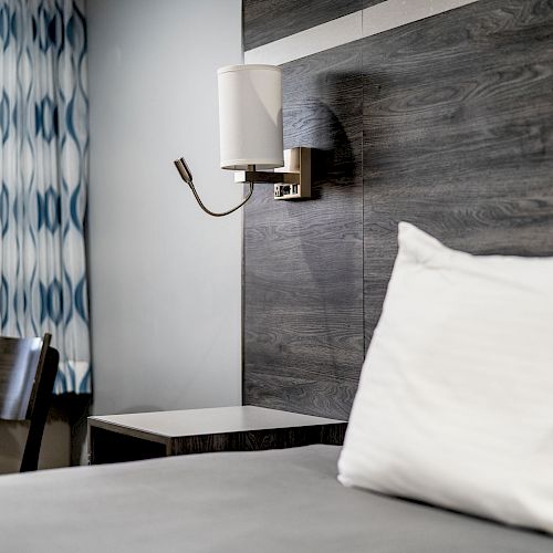 The image shows a modern hotel room with a neatly made bed, white pillows, a wall-mounted lamp, a wooden headboard, and a window with patterned curtains.