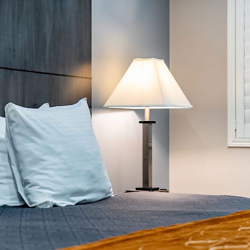 A cozy bedroom with neatly arranged white pillows on a dark bedspread, a lit bedside lamp, and a window with white shutters.