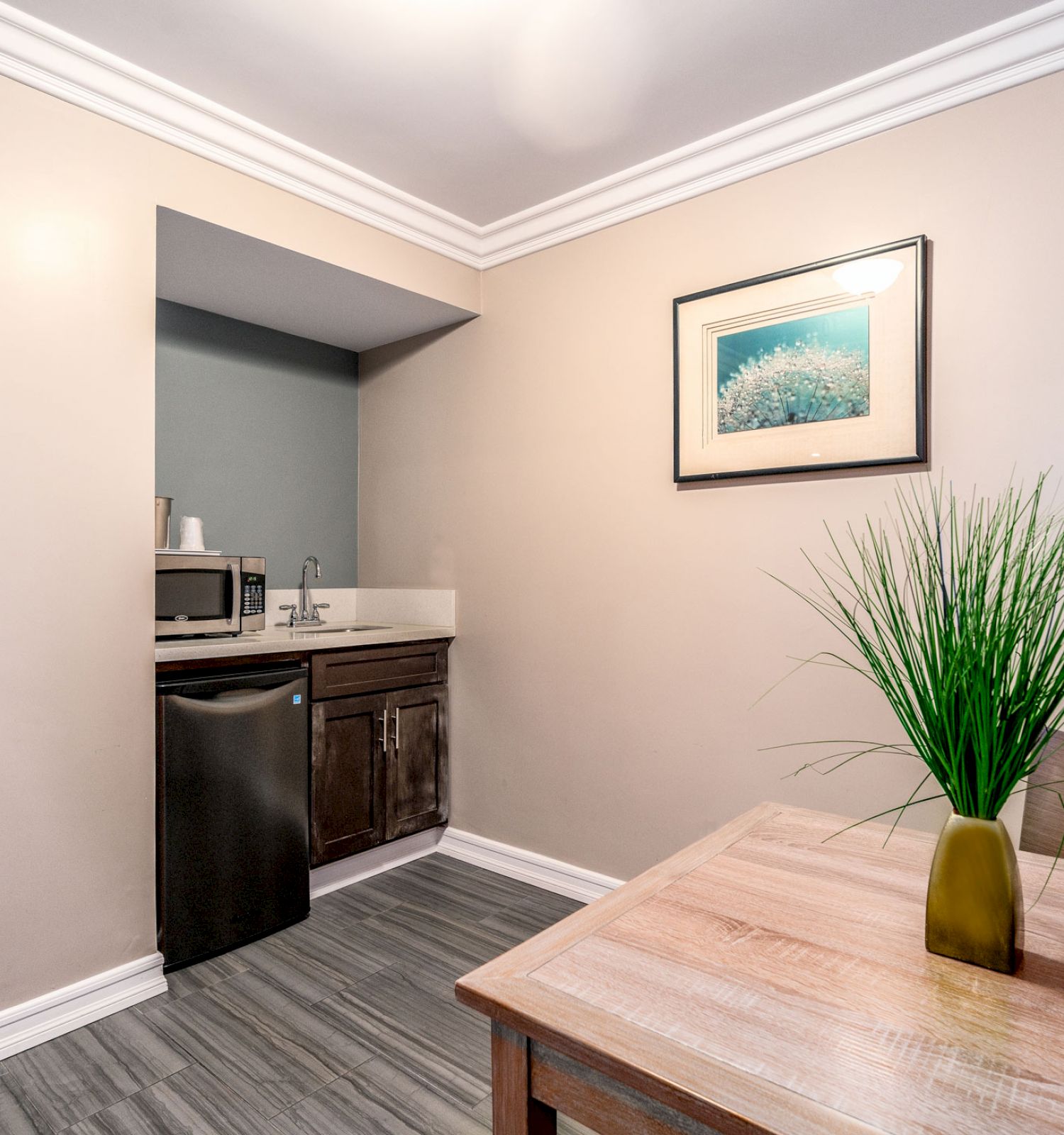 This image shows a small kitchen nook with lightwood cabinetry, a microwave, a mini-fridge, and a sink. Art pieces and a plant decorate the space.