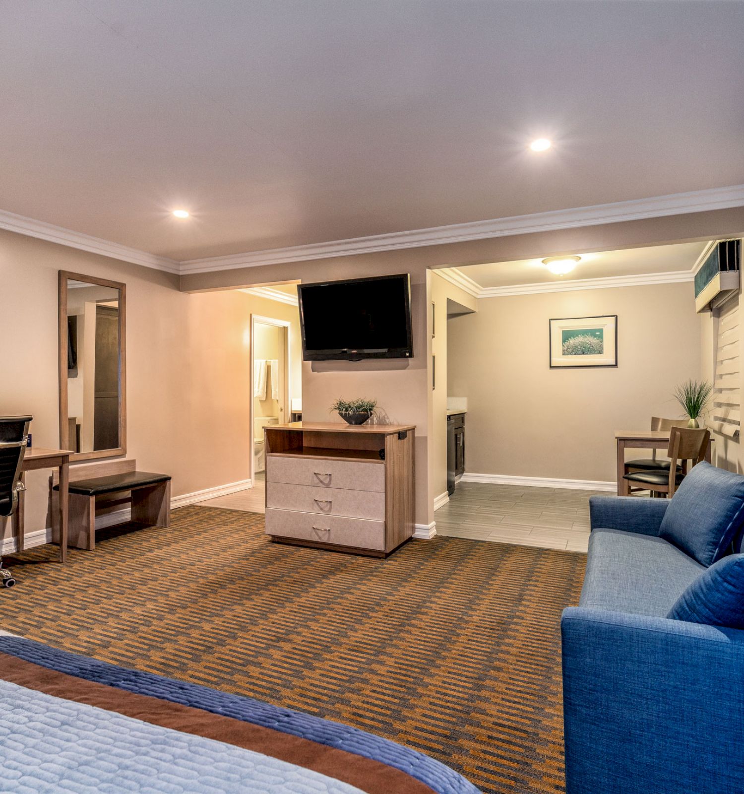 A spacious hotel room with a bed, sofa, TV, and workspace. In the background, a dining area is visible with a table and chairs.