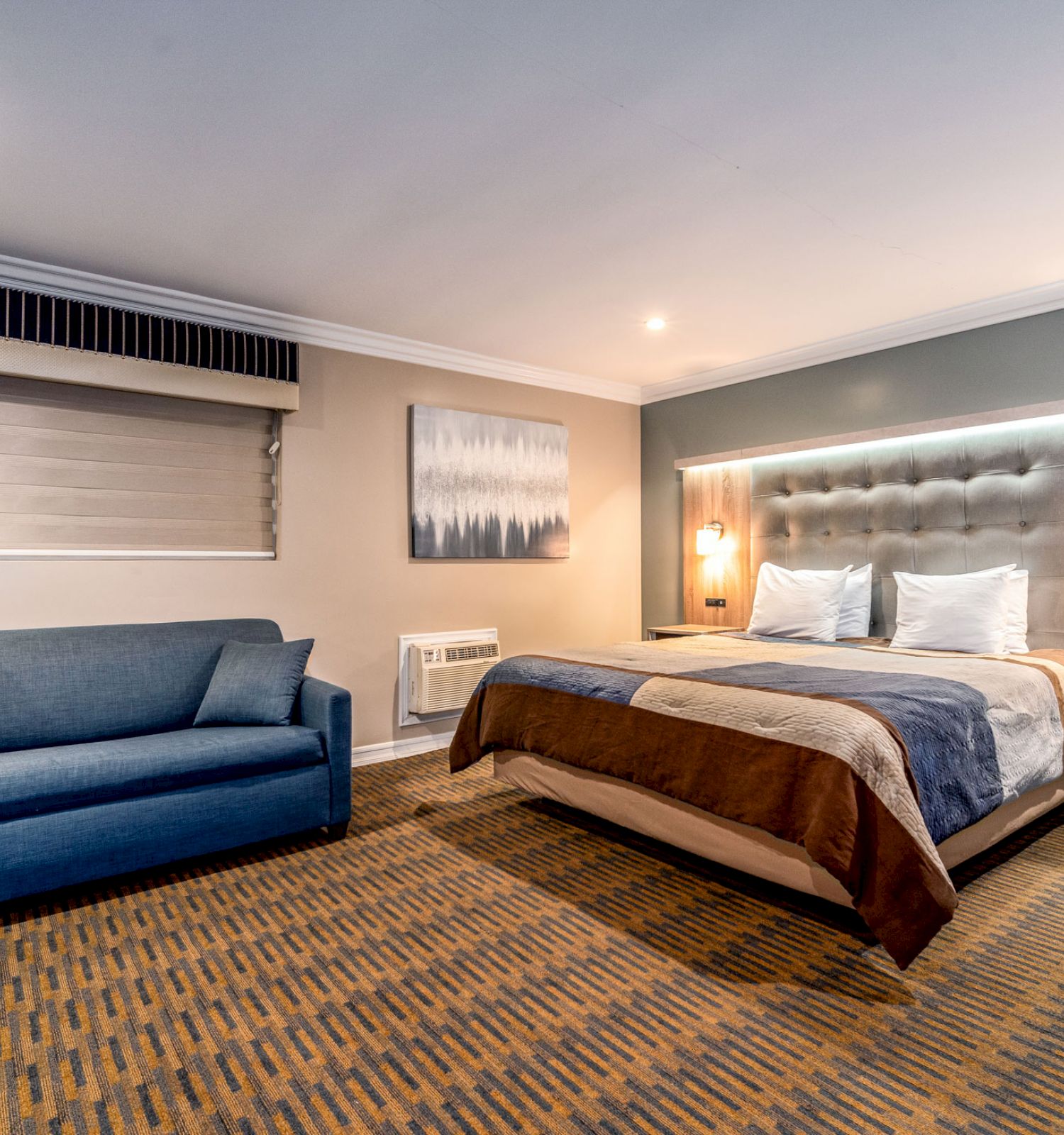 A modern hotel room with a double bed, upholstered headboard, bedside tables, a blue sofa, wall art, and carpeted flooring, well-lit.