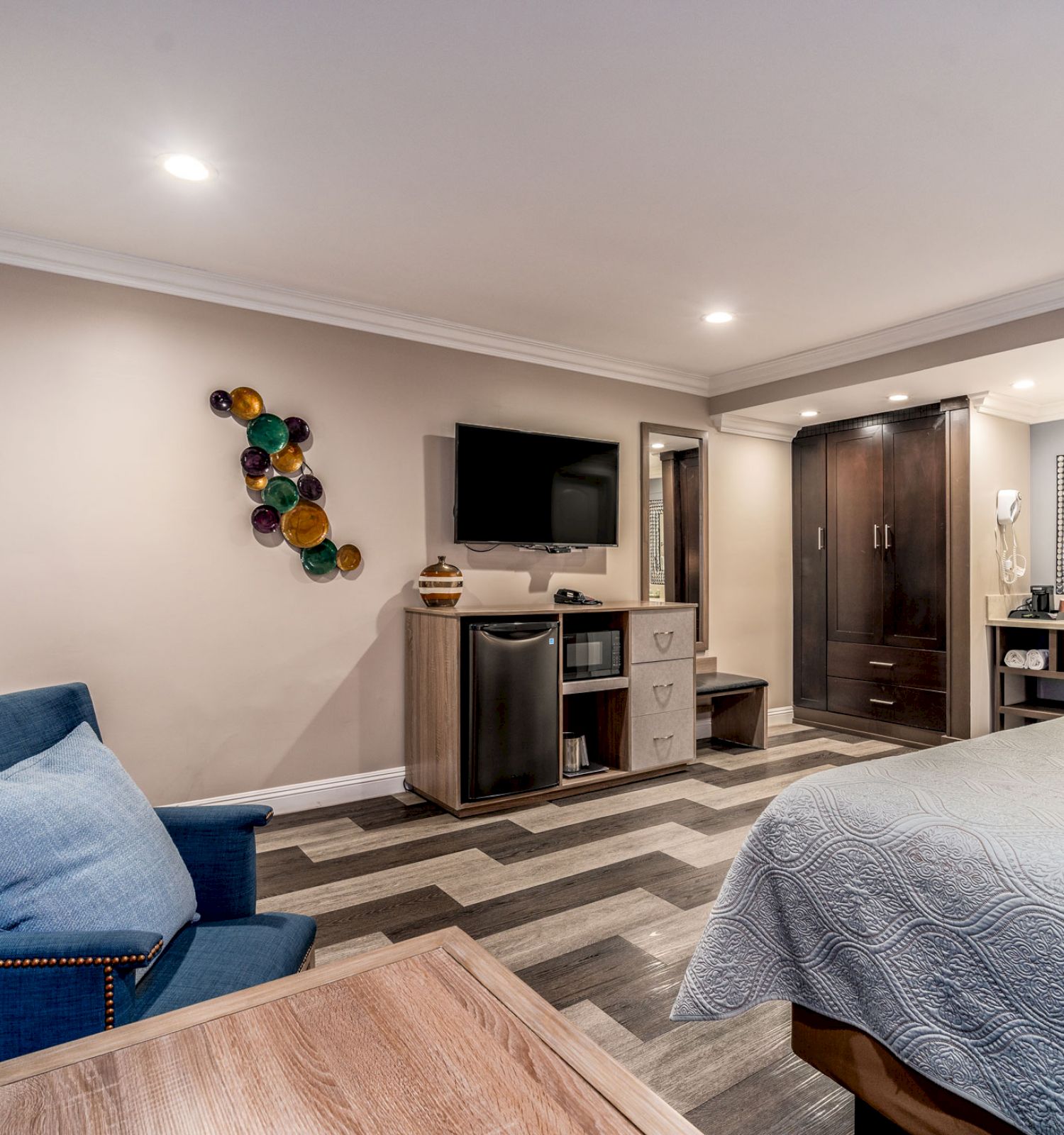 A modern bedroom with a double bed, a blue armchair, a TV mounted on the wall, a small desk, and a bathroom area.