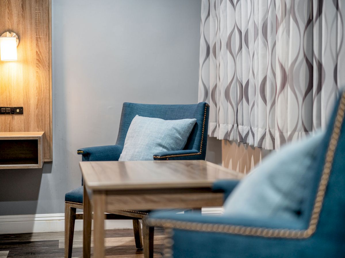 Two blue chairs with cushions, a wooden table, patterned curtains, and a wall-mounted light fixture in a cozy, softly lit room.