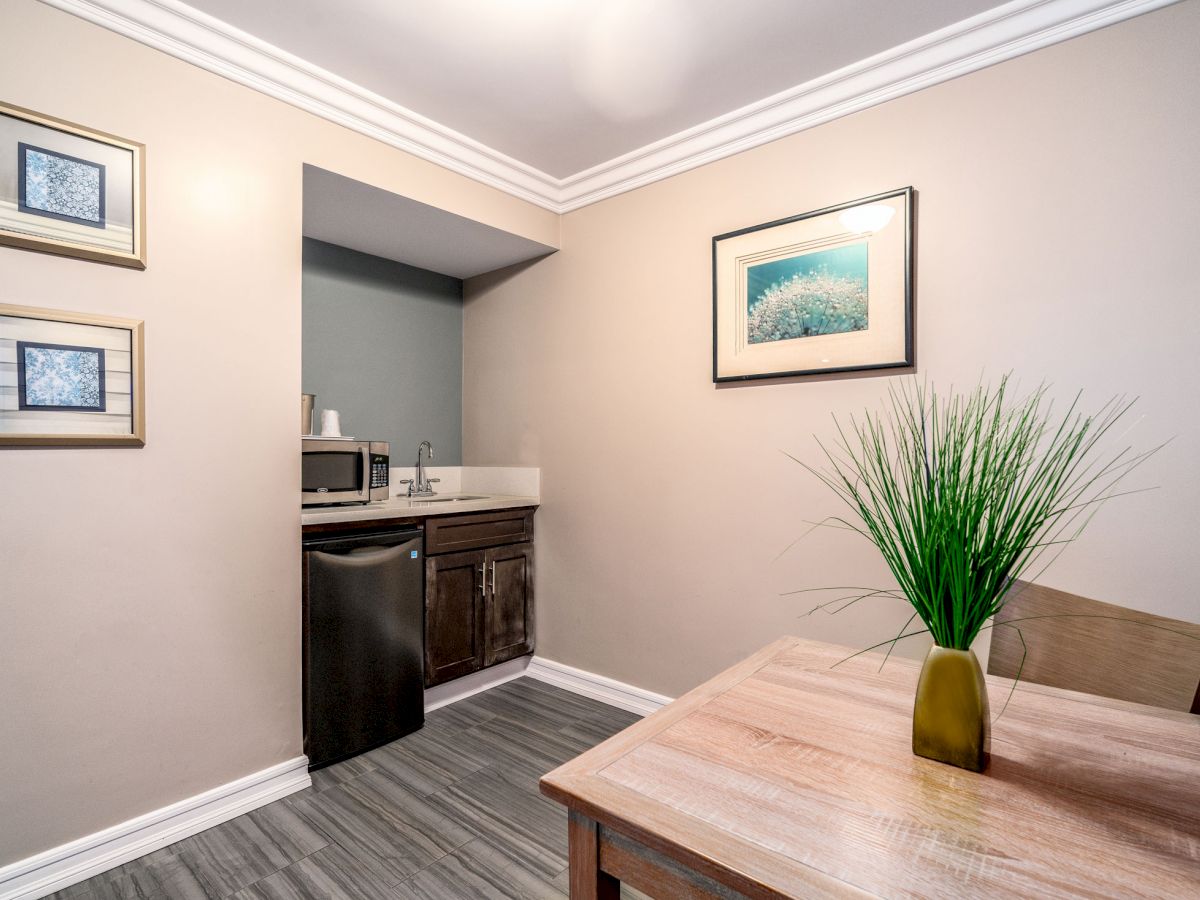 A small, modern kitchenette with a microwave, sink, and mini-fridge. There's a wooden table, chairs, framed artwork, and decorative plant.