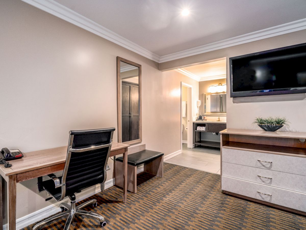 A hotel room workspace with a desk, chair, phone, mirror, TV, dresser, and a view of a bathroom; decorated in neutral tones and modern style.