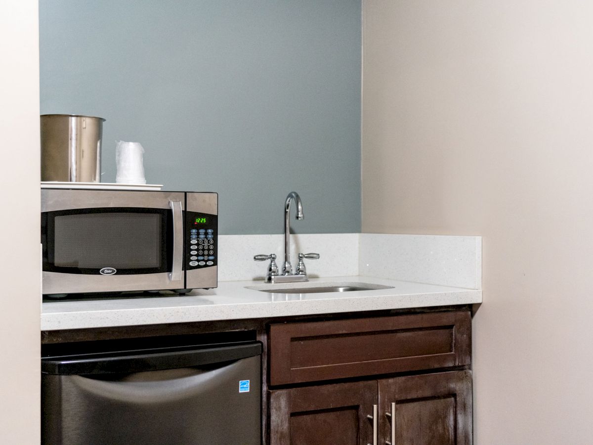 The image shows a small kitchenette with a microwave, sink, refrigerator, and storage cabinets. There’s also a metal ice bucket and some paper cups on the countertop.