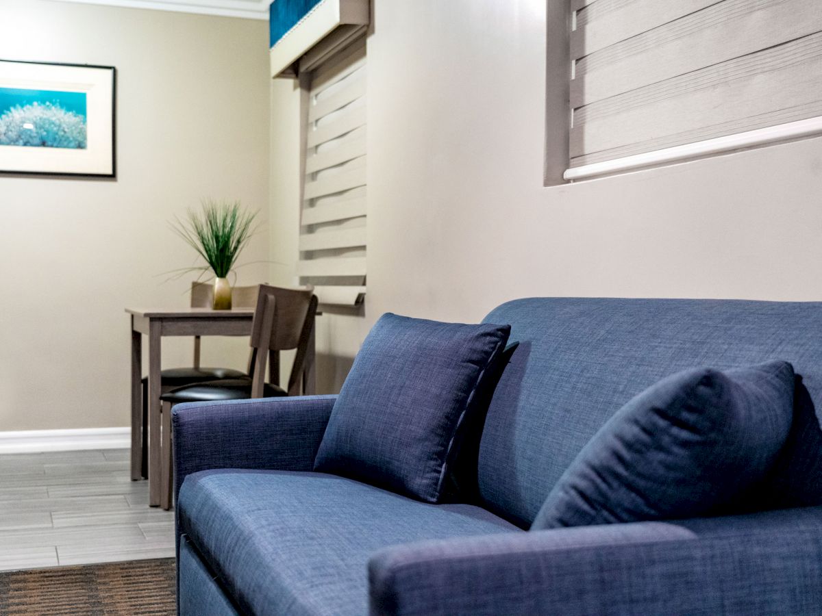 A room with a blue couch, a small wooden table with a chair, a plant, and a framed picture on the wall. The window has horizontal blinds.