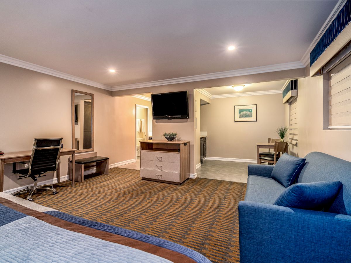 A well-lit hotel room features a bed, sofa, TV, desk, and seating area with modern decor.