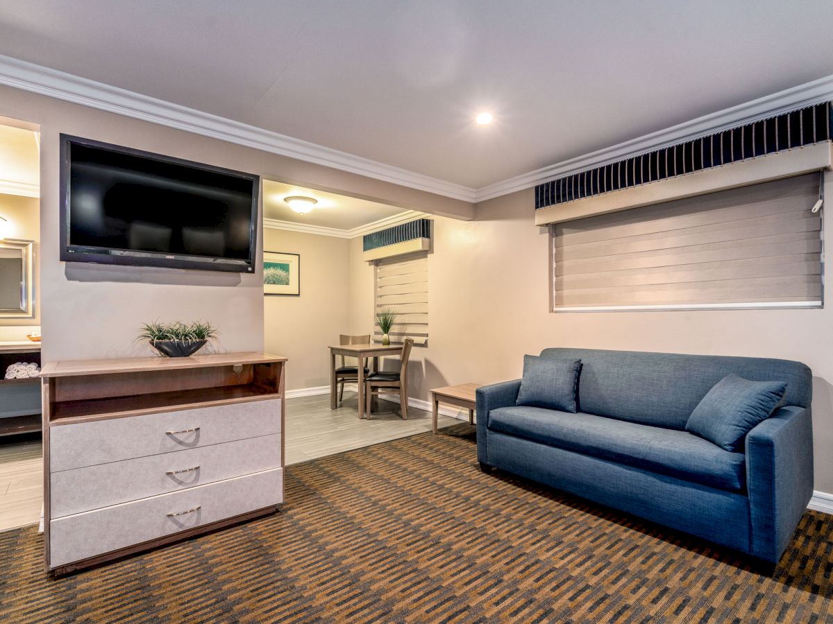 The image shows a hotel room with a blue couch, wall-mounted TV, dresser, and a small dining area in the background, featuring a cozy ambiance.