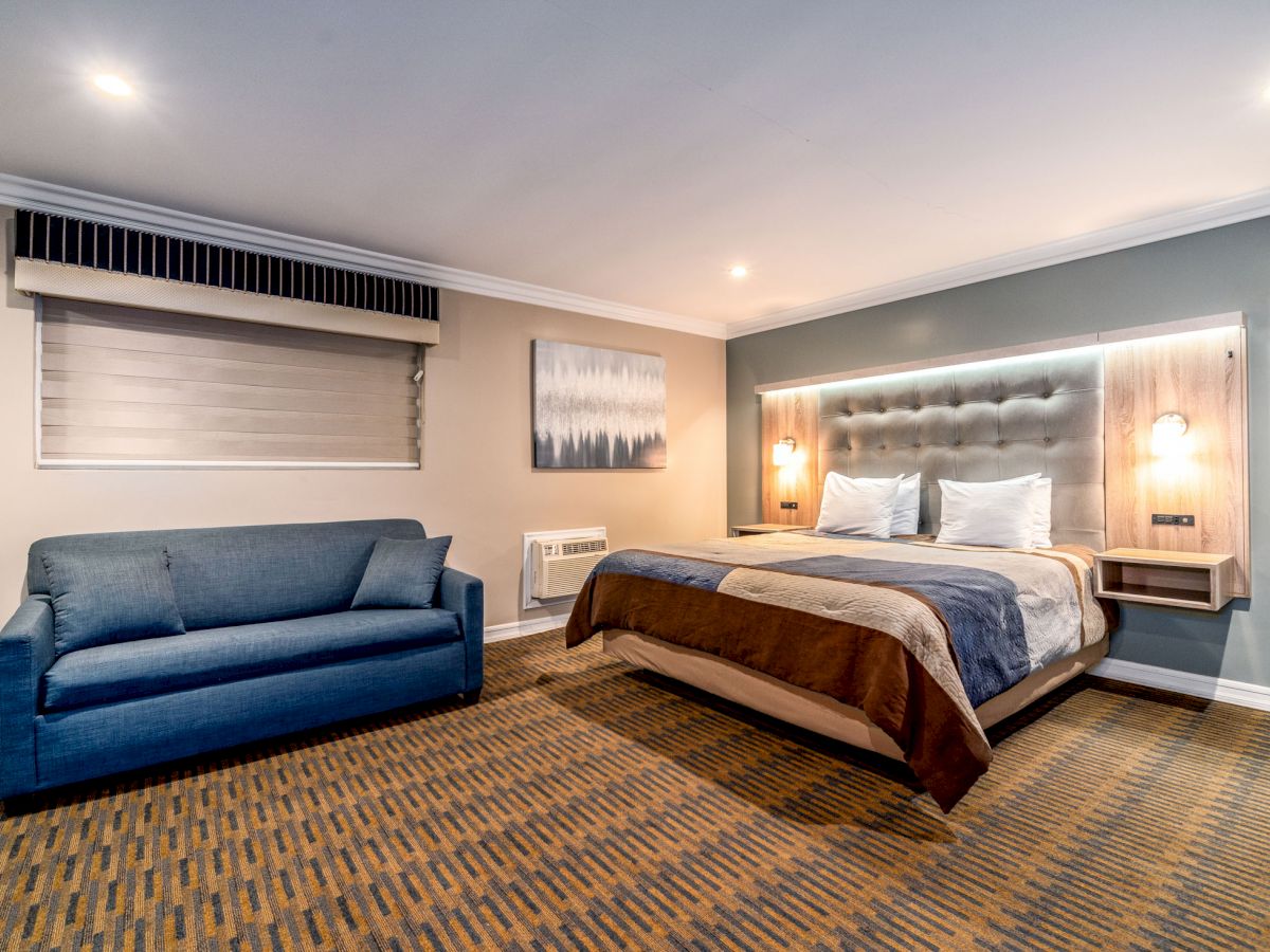 The image shows a modern hotel room with a bed, matching nightstands, a blue sofa, a painting, overhead lighting, and patterned carpet flooring.