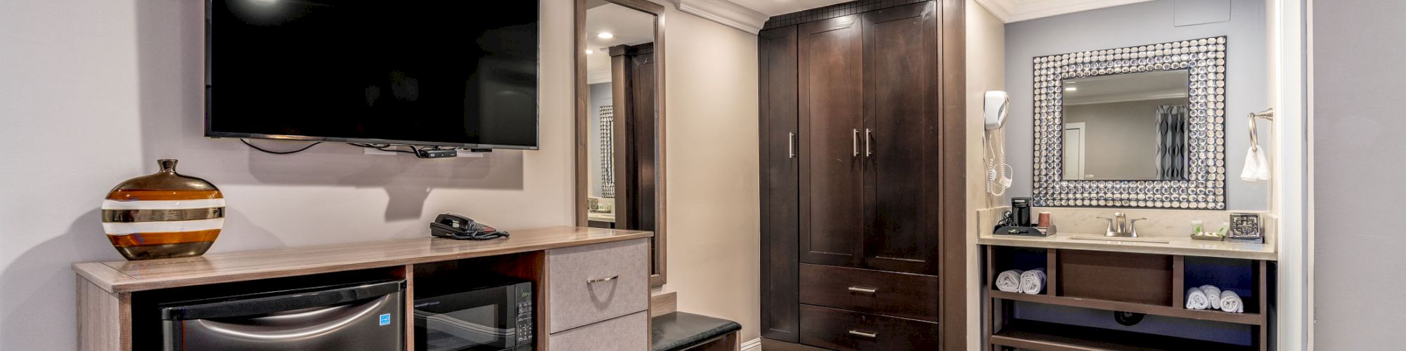 The image shows a modern hotel room with a TV, mini-fridge, a wardrobe, a mirror, and a bench, with wooden flooring and a neutral color scheme.