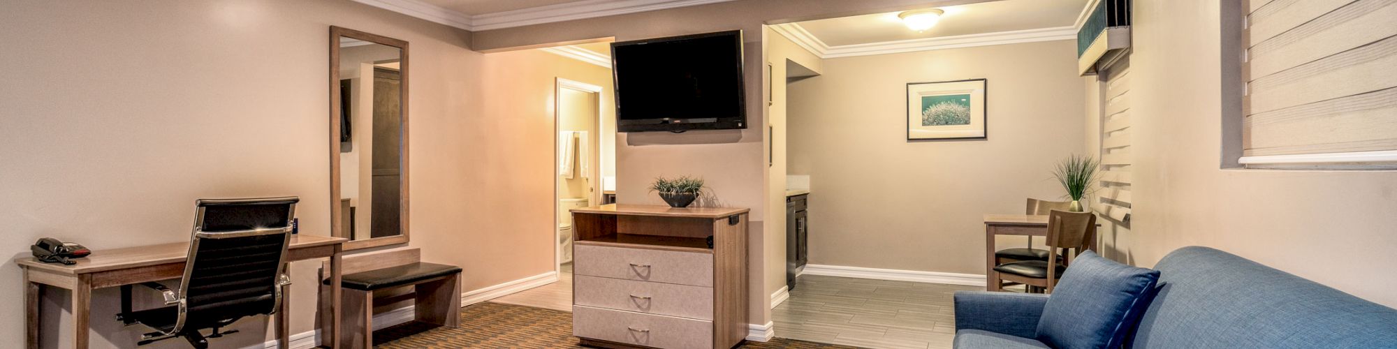 A modern hotel room with a bed, sofa, desk, TV, and artwork on the wall, featuring an open layout and neutral tones for a cozy atmosphere.