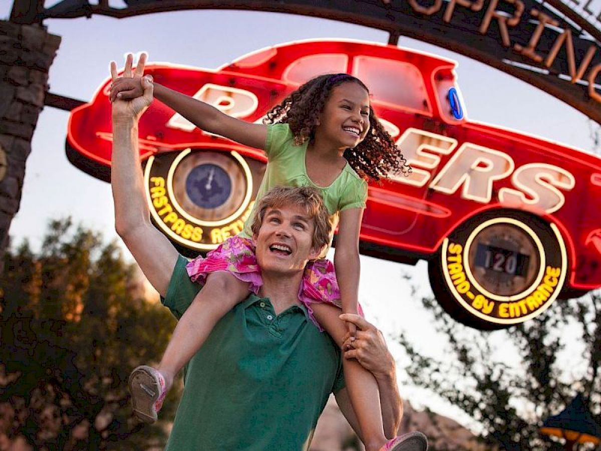 A man is carrying a smiling girl on his shoulders, standing in front of an amusement park sign featuring a red race car.