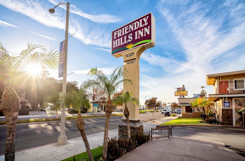 The image shows a street scene with a sign for "Friendly Hills Inn" against a bright, sunny sky and palm trees. The inn is located at 14329.