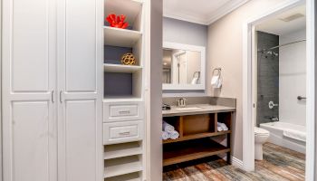 The image shows a modern bathroom with white cabinets and shelves, a large mirror above a sink with an open vanity, and a bathtub in the adjacent room.