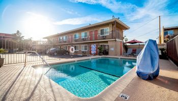 An outdoor swimming pool area with a protective cover, adjacent to a two-story motel building with a sunny sky above and parked cars in the background.