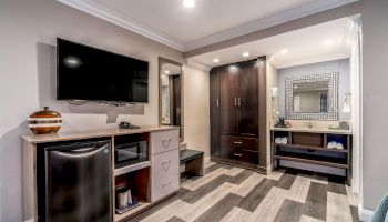 A modern hotel room features a TV, mini-fridge, drawers, large wardrobe, and vanity area with a mirror and shelves.