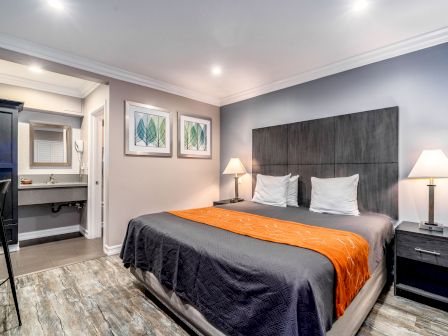 A modern bedroom with a double bed, orange runner, side tables, lamps, wall art, and an open door to a bathroom in the background.