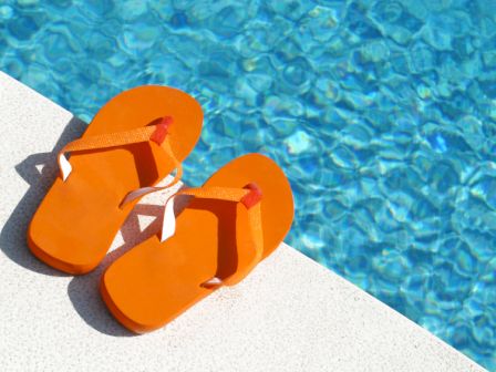 The image shows a pair of orange flip-flops on the poolside edge next to a clear blue swimming pool.