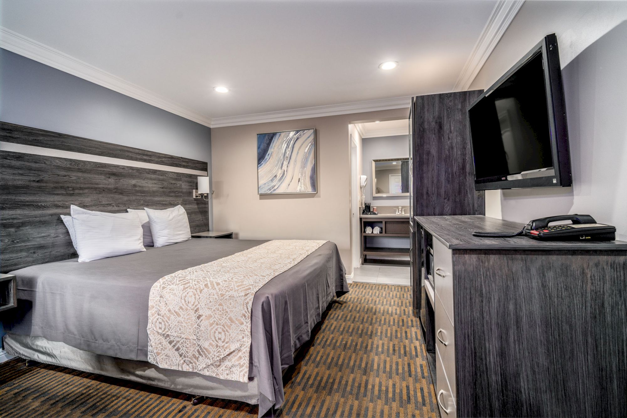 A modern hotel room with a large bed, TV, dresser, and an open bathroom door showing a sink and mirror in the background.