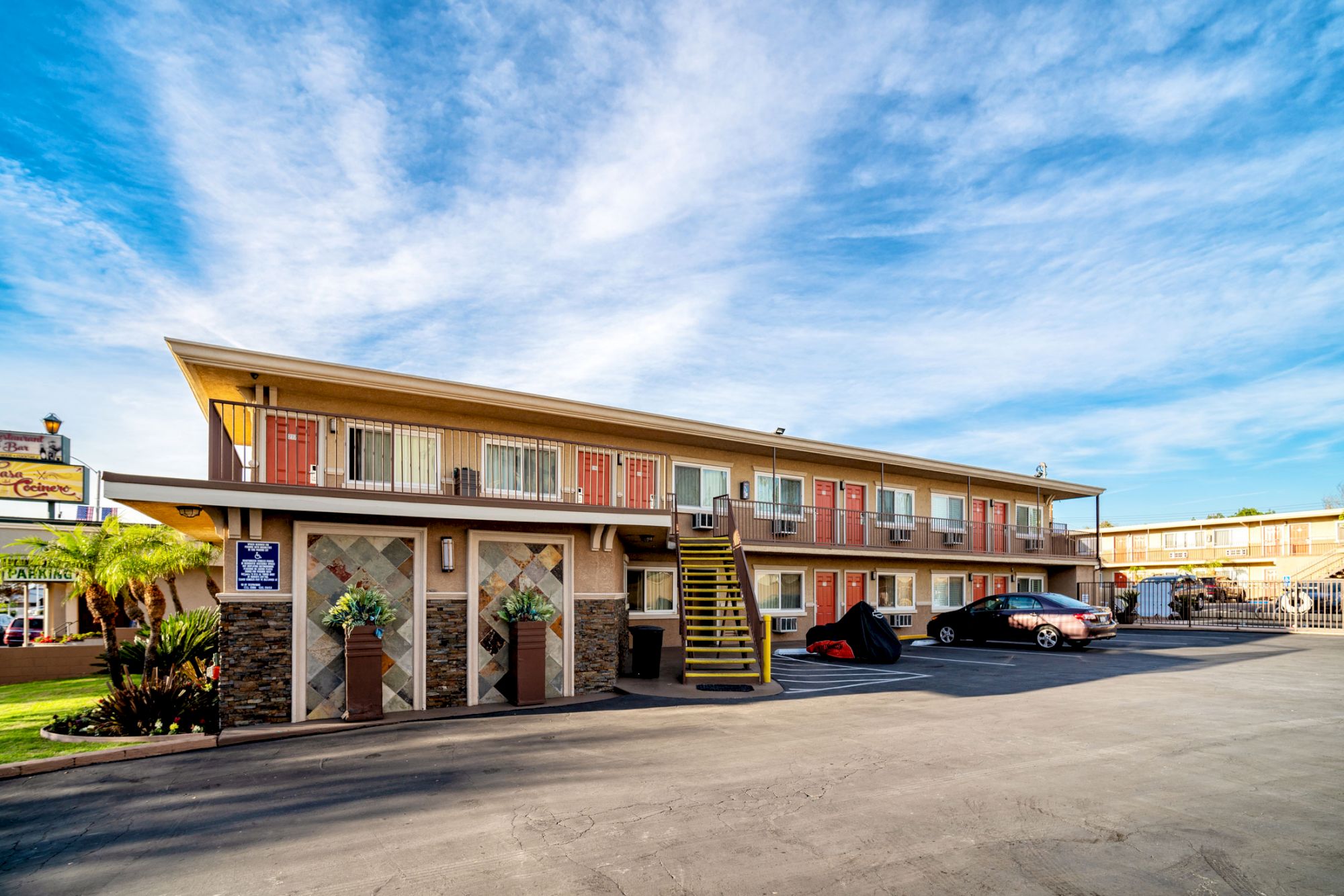 The image shows a two-story motel with external staircases and parking spaces in front, under a clear blue sky with a few clouds.