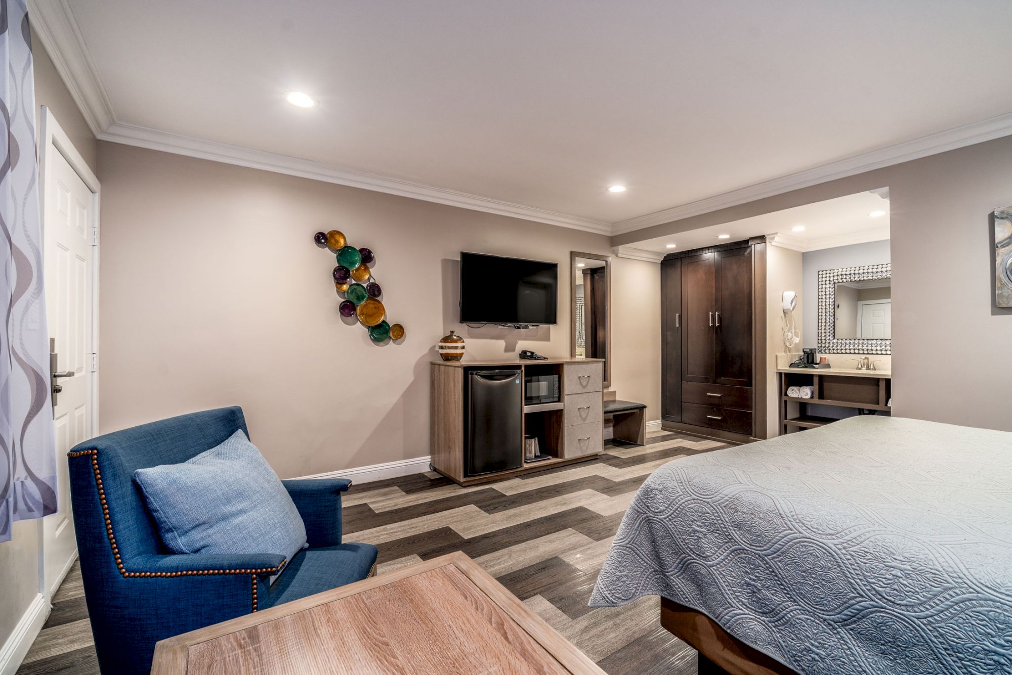 The image shows a modern hotel room with a bed, a blue chair, a flat-screen TV, wooden floors, and a separate vanity area with cabinets.